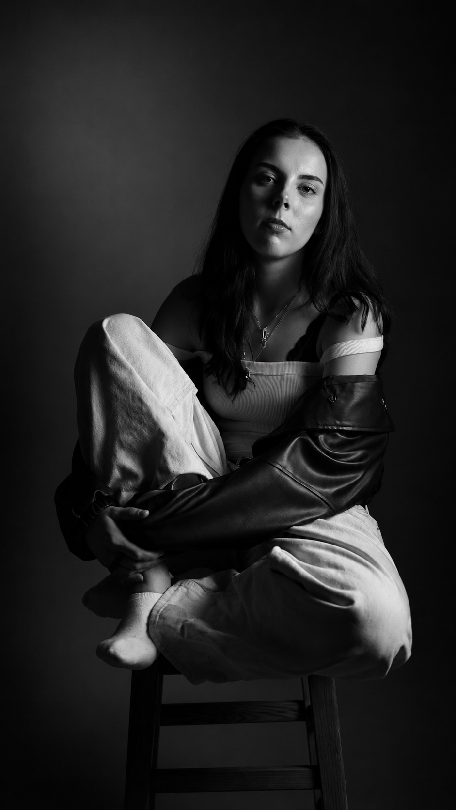 A young woman in a leather jacket sitting cross-legged on a stool against a dark studio backdrop