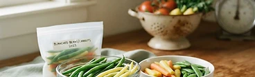 A variety of colorful, blanched vegetables including green beans and peas cooling in a stainless steel colander next to a bowl of ice water.