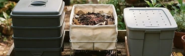 Three different types of worm bins sitting on a wooden deck next to a lush vegetable garden.