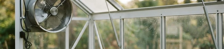 A modern hobby greenhouse with open roof vents and a small oscillating fan visible inside among green vegetable plants.