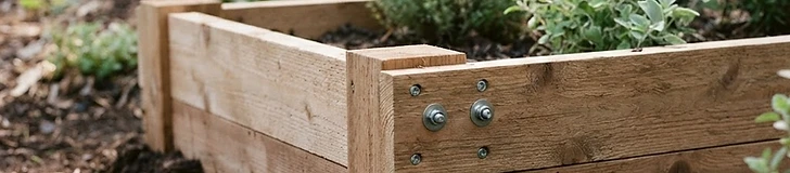 A close-up shot of a pair of hands using a cordless drill to secure a cedar board to a corner post of a raised garden bed.