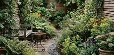 A small urban courtyard garden featuring vertical wooden planters on a brick wall, a bistro set on a light gravel path, and layered green foliage.