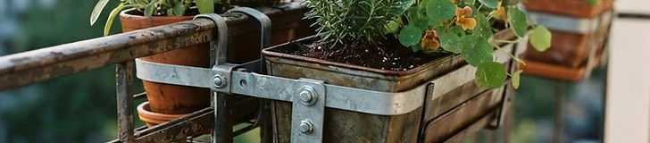 A close-up view of a metal railing planter securely attached to a balcony guardrail with visible heavy-duty brackets and lush green herbs growing inside.