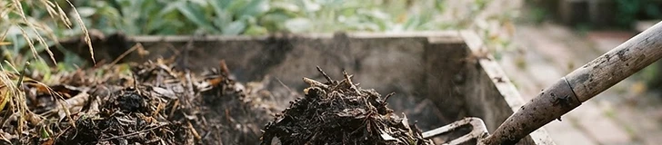 A gardener using a metal pitchfork to turn a wooden compost bin filled with a mix of dried brown leaves and fresh green vegetable scraps.