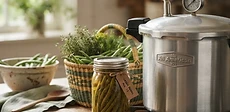 A stainless steel pressure canner sitting on a stovetop surrounded by glass mason jars filled with fresh green beans and carrots.
