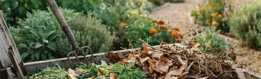 A close-up view of a garden compost pile showing a mix of dry brown leaves and fresh green vegetable scraps being turned with a pitchfork.
