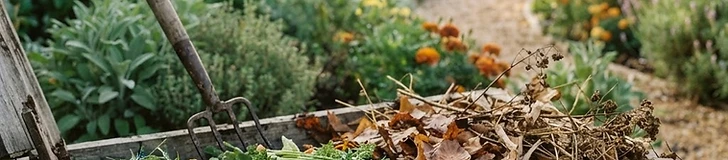 A close-up view of a garden compost pile showing a mix of dry brown leaves and fresh green vegetable scraps being turned with a pitchfork.