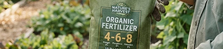 A close-up view of a gardener's hands holding a green fertilizer bag with the N-P-K numbers clearly printed on the label, with blurred garden rows in the background.