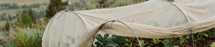 A raised garden bed covered with white floating row covers on a sunny mountain hillside, with pine trees visible in the background.