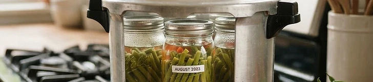 A stainless steel pressure canner sitting on a clean kitchen stovetop with several glass canning jars filled with green beans nearby.