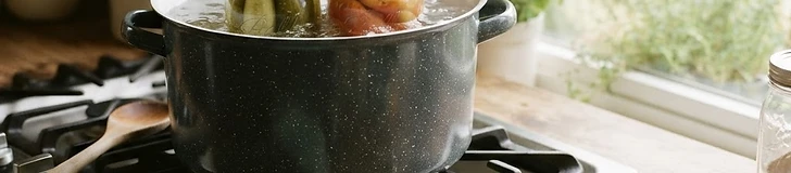 Close-up of glass mason jars filled with bright red tomatoes and sliced peaches sitting on a rustic wooden table next to a boiling water-bath canner.