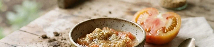 Close up of a gardener scooping tomato seeds and pulp into a small glass jar on a wooden potting bench.