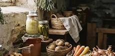 A rustic wooden basket filled with freshly harvested onions, carrots, and winter squash sitting on a wooden table in a sunny garden shed.