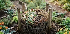 A close-up view of a gardener's hands placing kitchen scraps into a wire-mesh compost basket centered in a stone-walled raised keyhole garden bed.