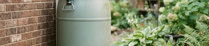 A blue plastic rain barrel positioned next to a brick house wall connected to a white metal downspout with a diverter hose.