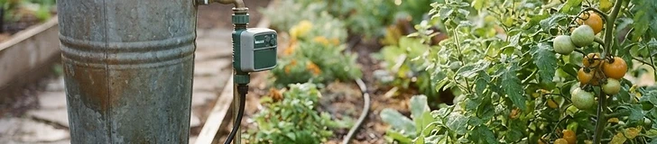 A blue plastic rain barrel positioned under a white house gutter downspout with a garden hose attached at the base.