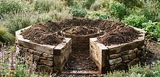 A circular raised garden bed made of stacked fieldstones with a wire mesh compost basket in the center and lush vegetable plants growing around it.
