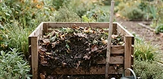 A backyard garden scene showing a plastic compost bin sitting on top of soil with a metal pitchfork leaning against it.
