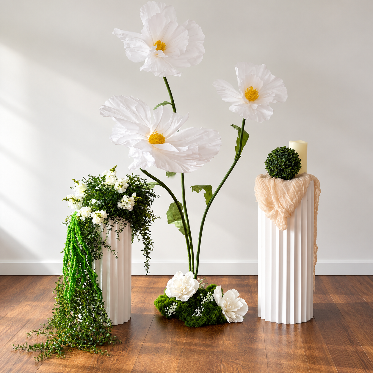 Studio display of oversized white paper flowers on white pedestals with trailing vines and a pillar candle