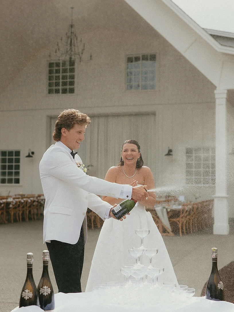A bride and groom popping champagne over a tower of coupes on a white-clothed table at an outdoor reception