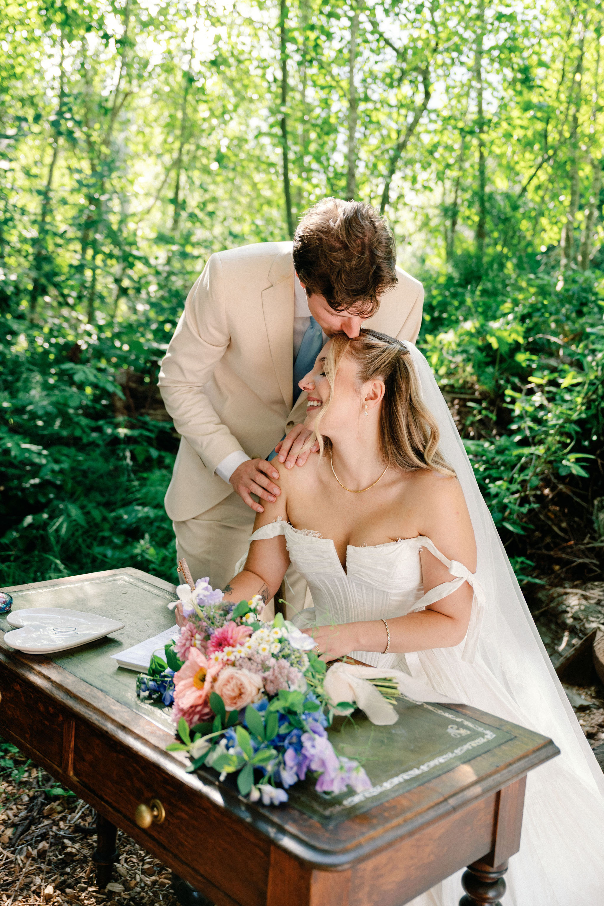 A bride seated at an antique desk in a sun-dappled forest as the groom tenderly kisses her forehead