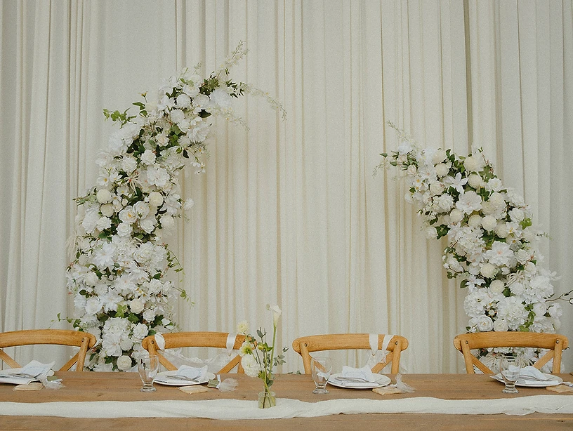 A wedding reception head table set against draped cream curtains, framed by lush white and cream floral arrangements, with cross-back chairs and place settings
