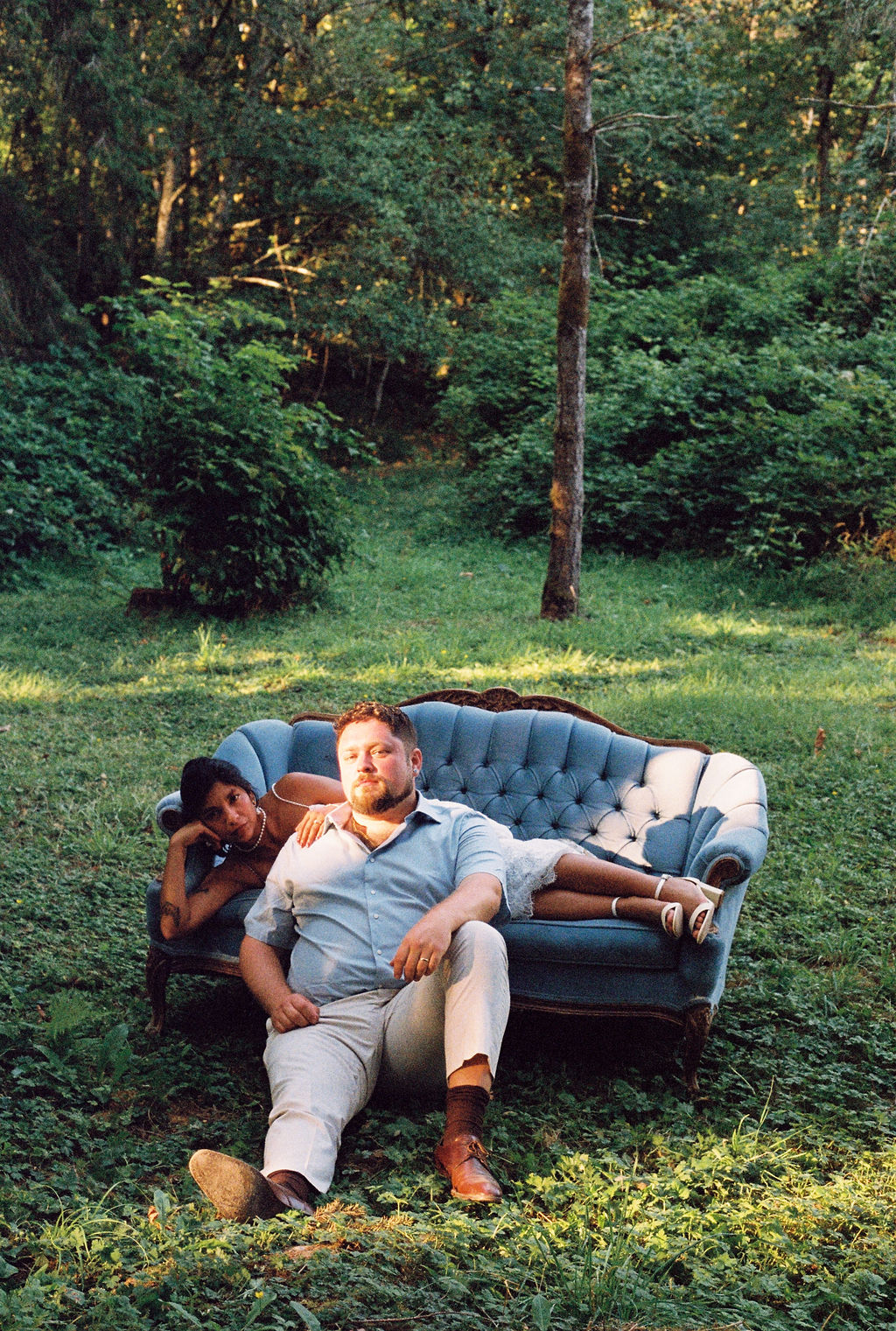 A couple posed on a vintage blue tufted sofa in a sunlit forest clearing
