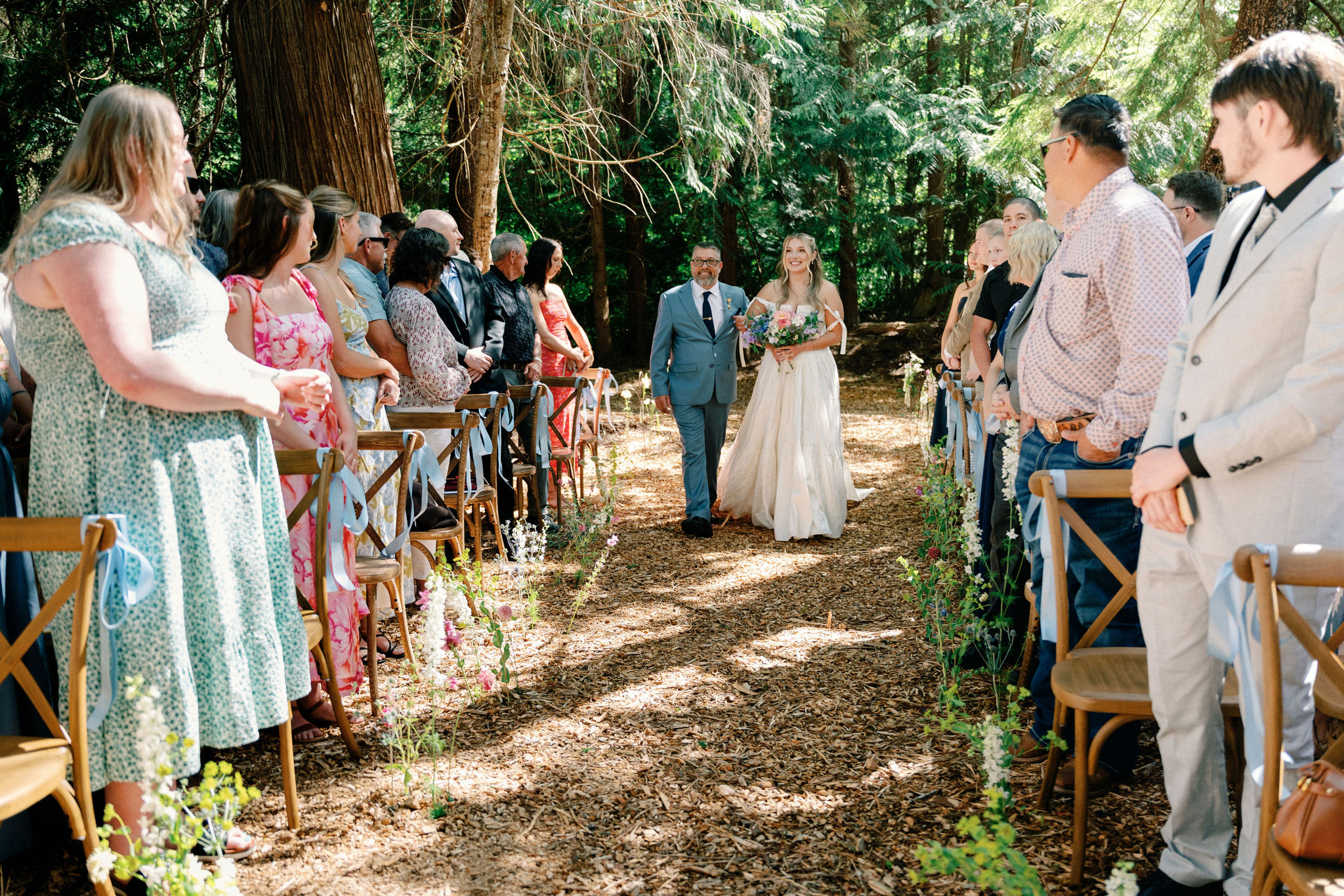 A bride walking down a woodland aisle escorted by a family member, guests seated on either side