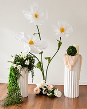Oversized white paper flowers on green stems between two fluted white pedestals styled with greenery, a topiary and a draped fabric