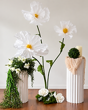Studio floral display with three oversized white paper flowers, a trailing ivy pedestal and a fabric-draped pedestal with topiary and candle