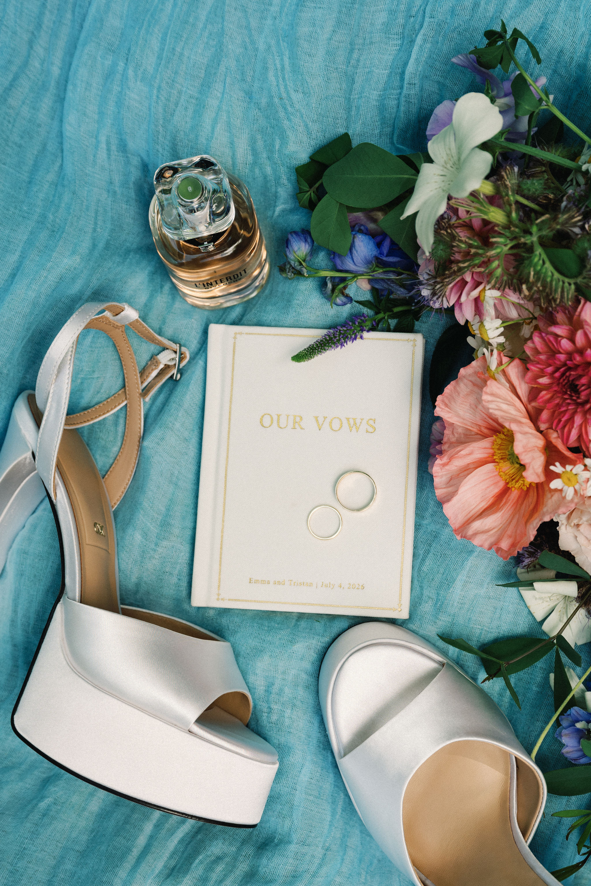 A flat lay of a vows booklet, wedding bands, satin heels, perfume and florals on a textured azure fabric