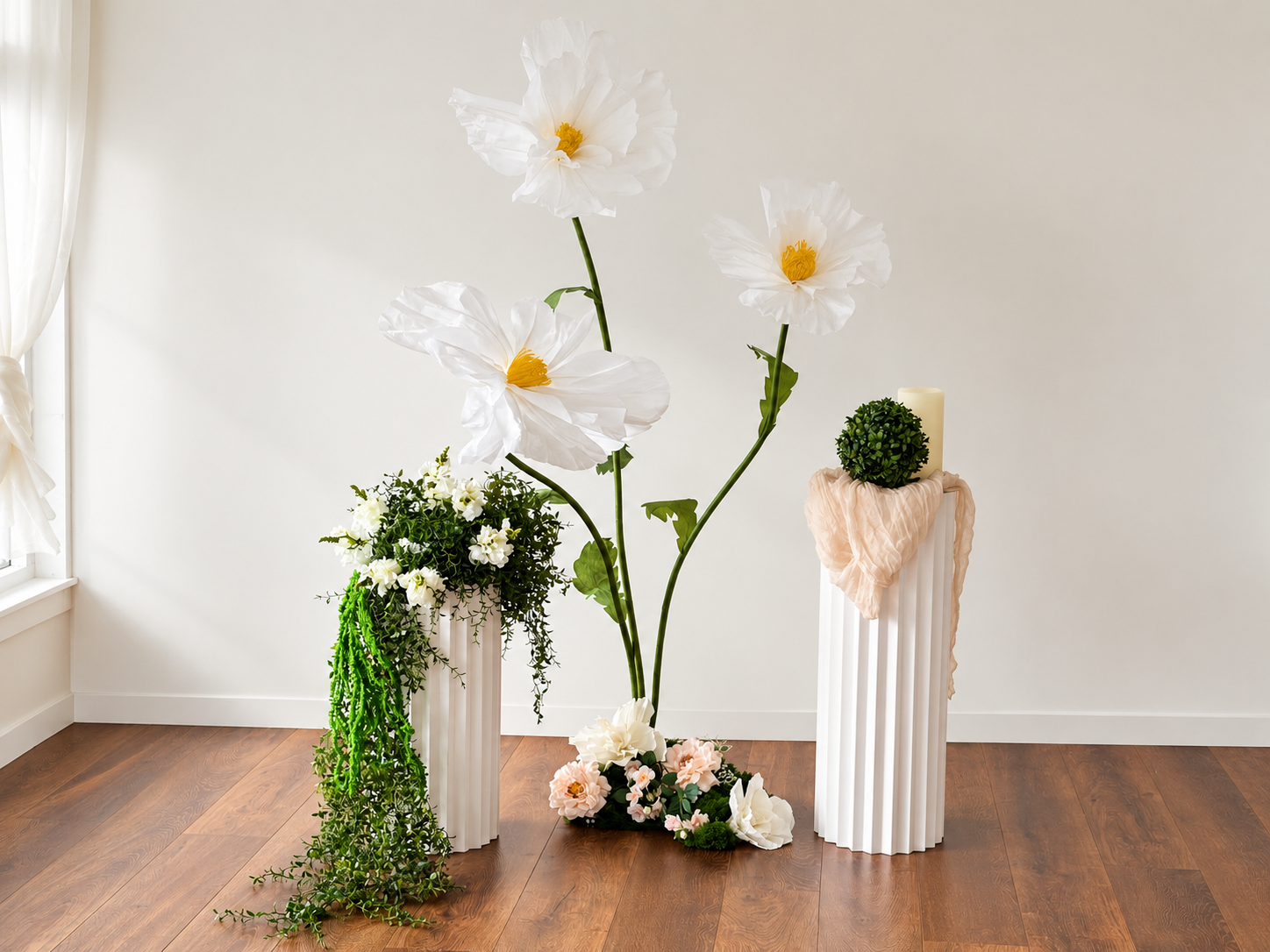 A minimalist event decor display with oversized white paper flowers, fluted pedestals, trailing greenery and a pillar candle