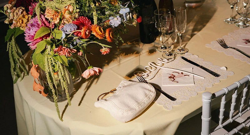 A wedding reception table setting with a vibrant floral centerpiece, beaded handbag and lace placemat in warm golden-hour light