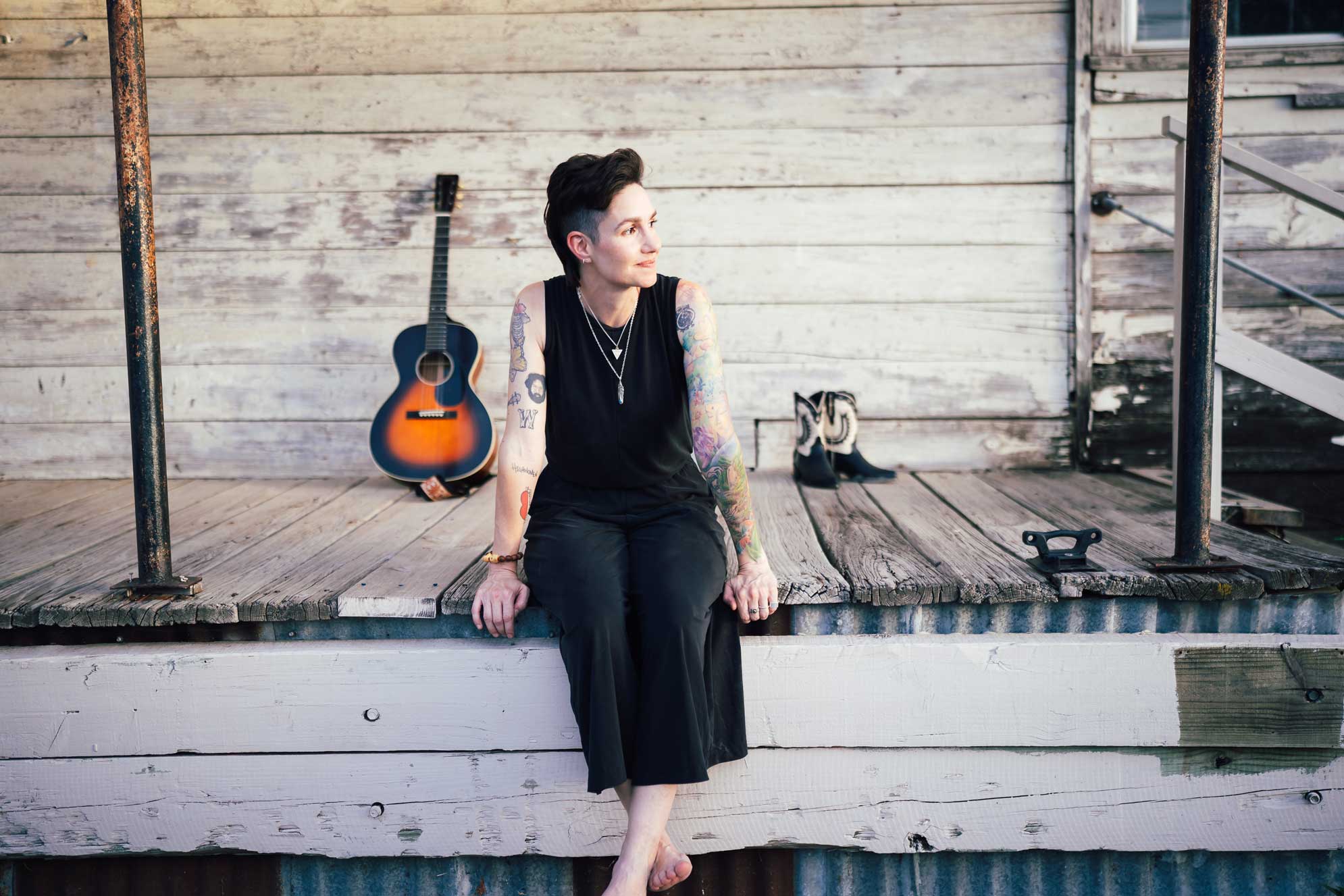 Emily McLoud seated barefoot on a weathered wooden porch edge with a sunburst acoustic guitar and cowboy boots, colorful tattoos, golden hour, Americana country folk artist