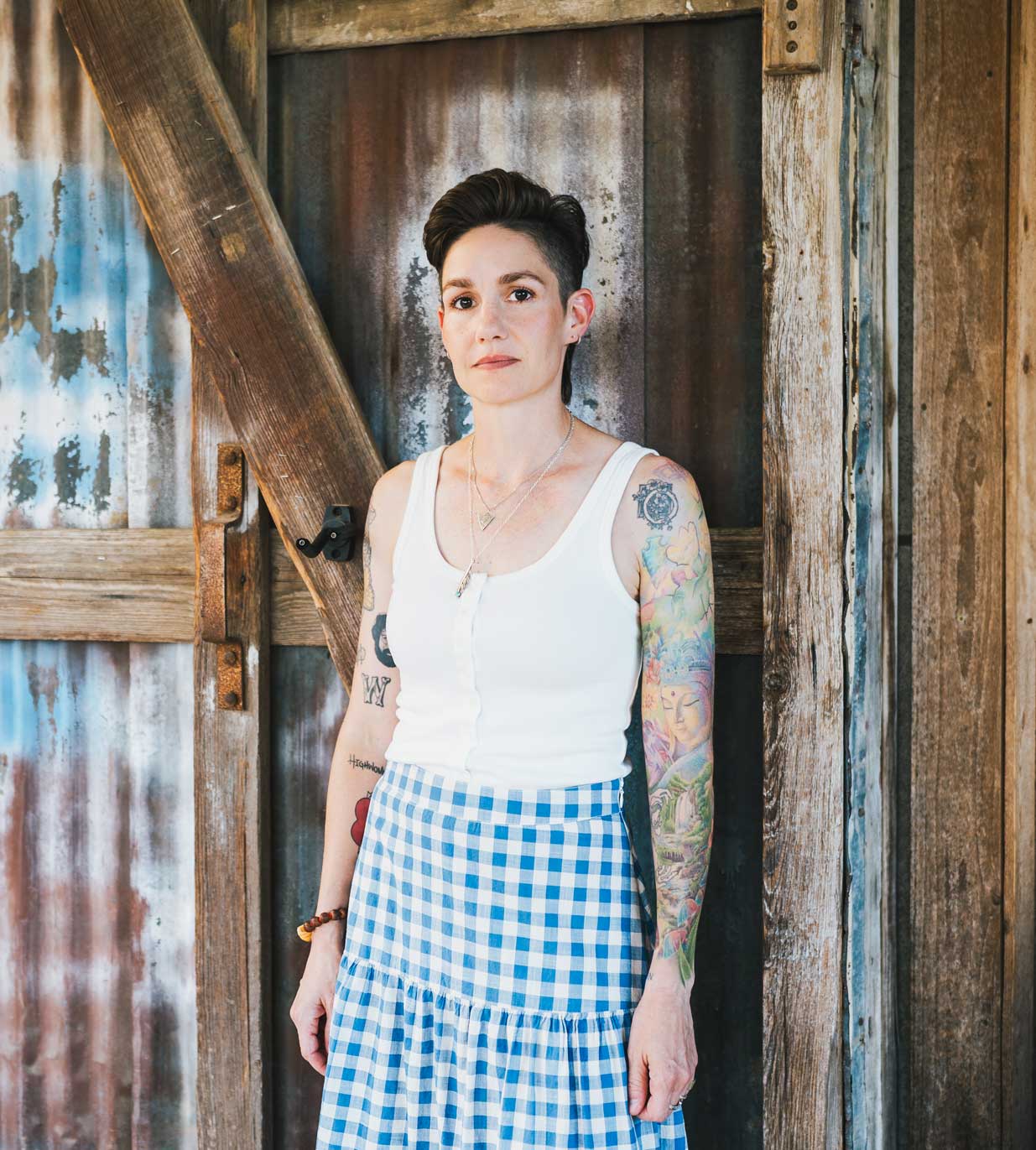 Emily McLoud standing at rustic barn door, white tank top and gingham skirt, tattoos
