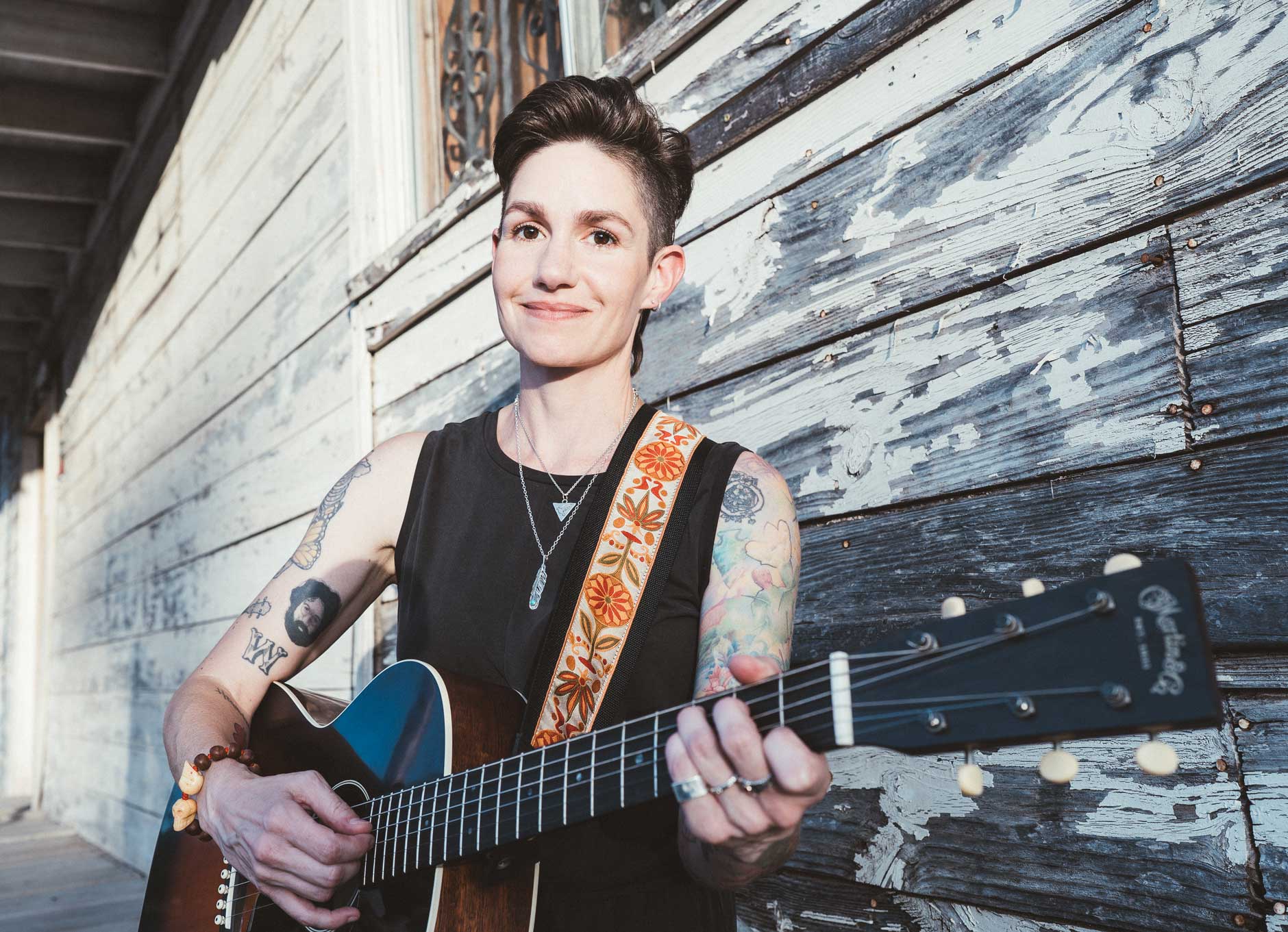 Emily McLoud with acoustic guitar against peeling white wood wall, soft light