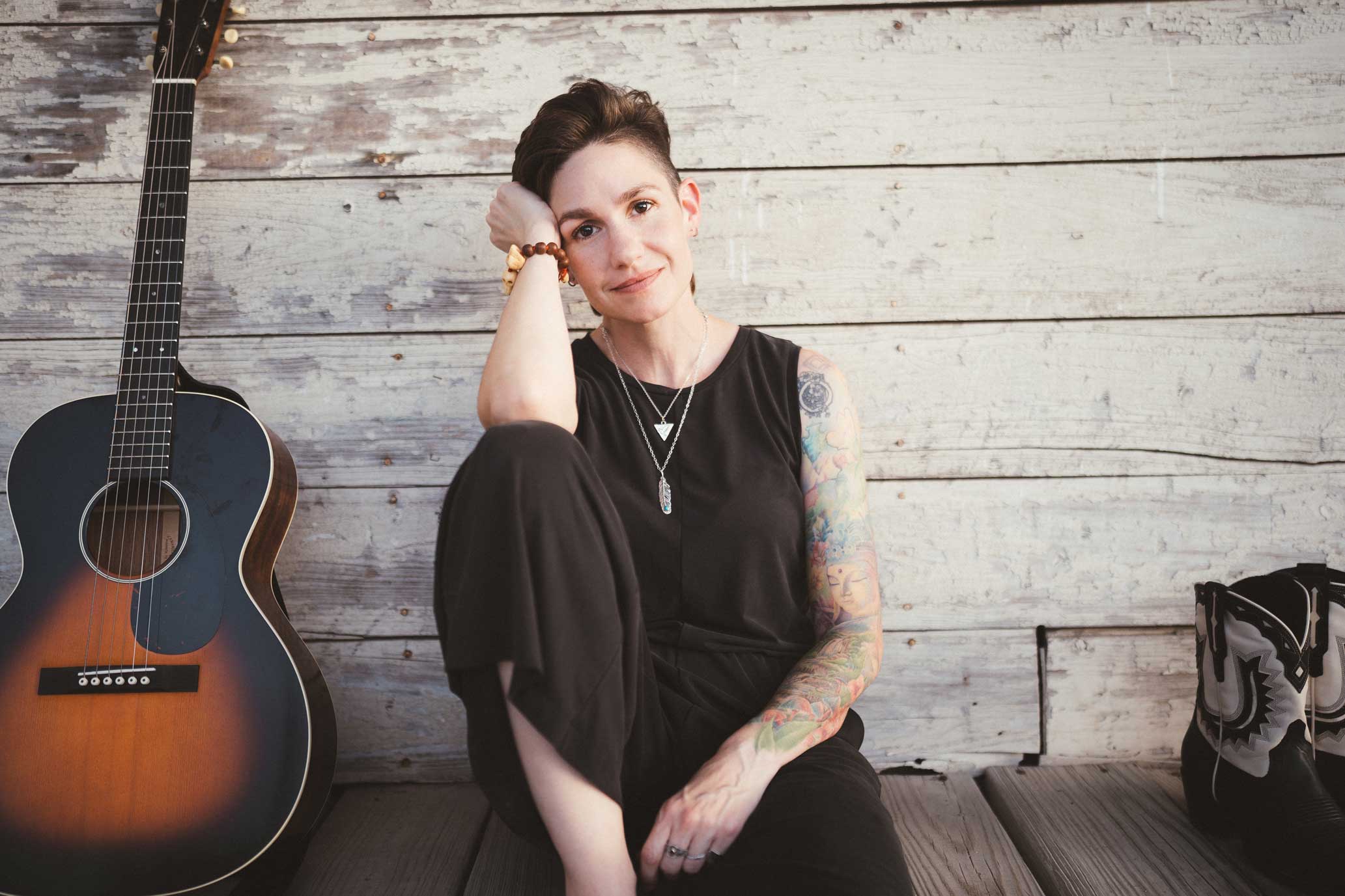 Emily McLoud seated with guitar and cowboy boots against white wood wall