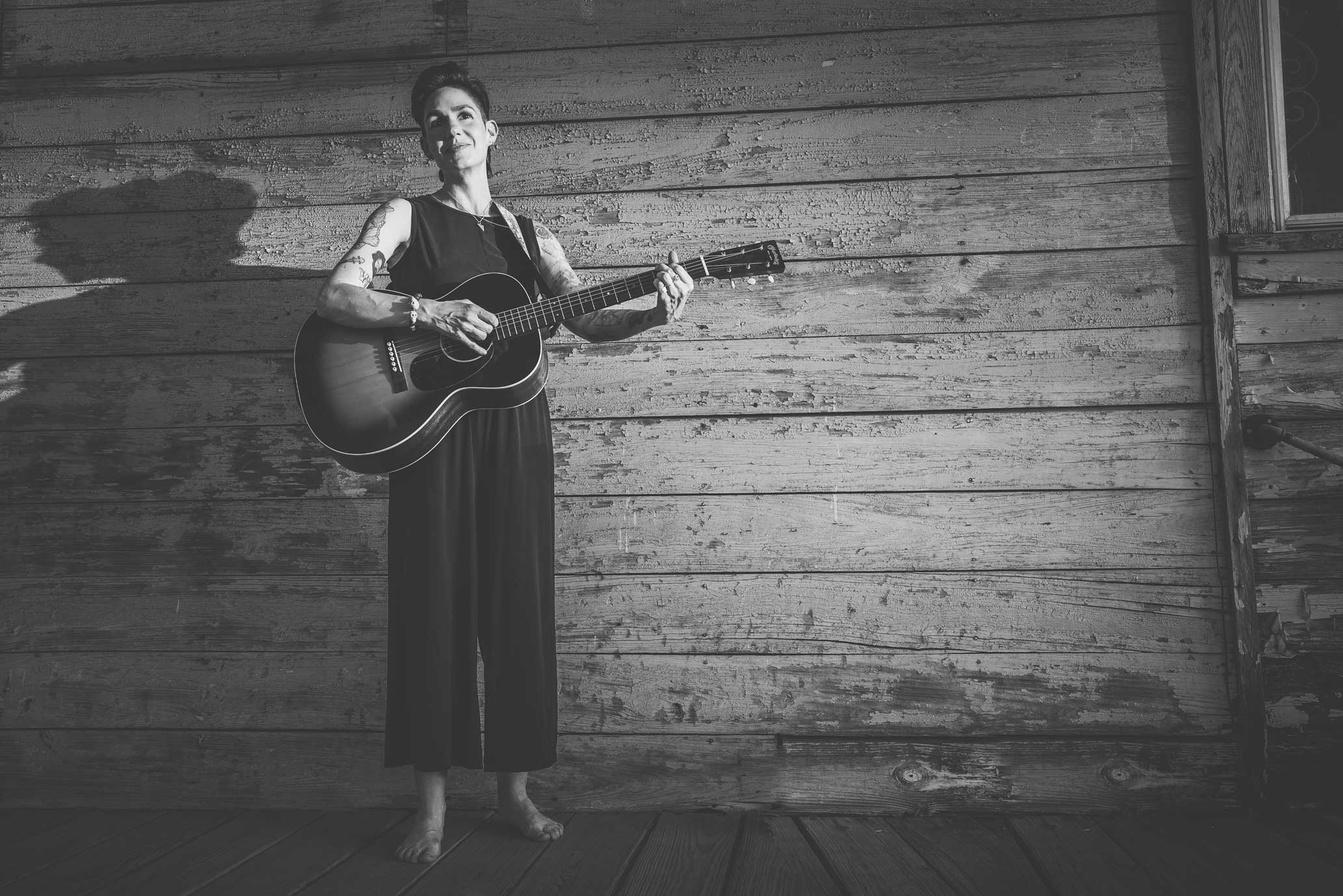 Emily McLoud black and white, full-length playing guitar standing against wood-plank exterior