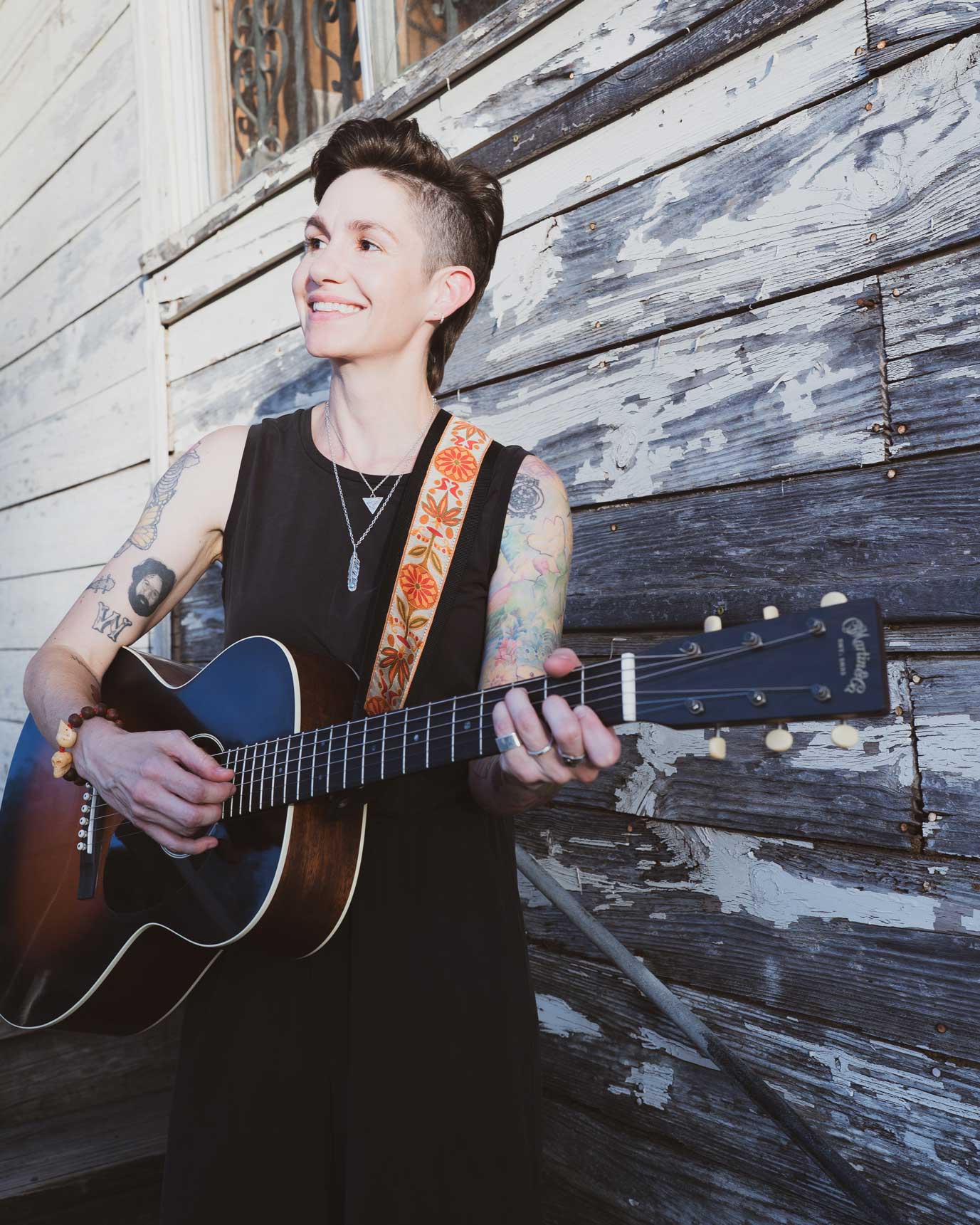 Emily McLoud playing guitar and smiling sideways, weathered exterior wall