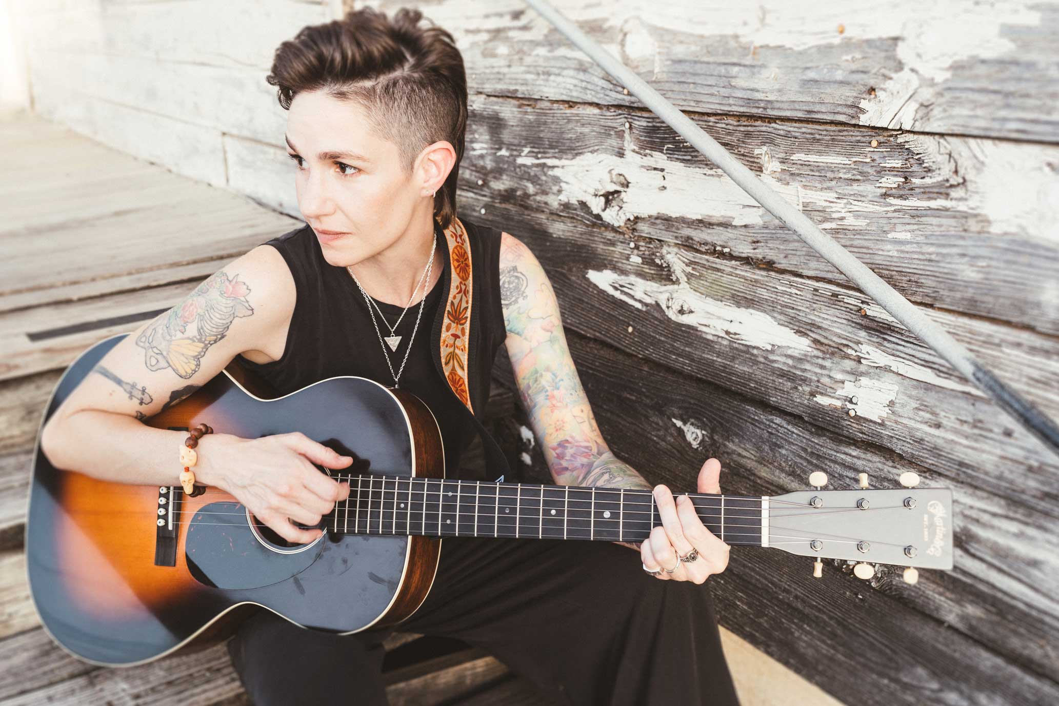 Emily McLoud seated playing acoustic guitar, looking off camera, rustic porch