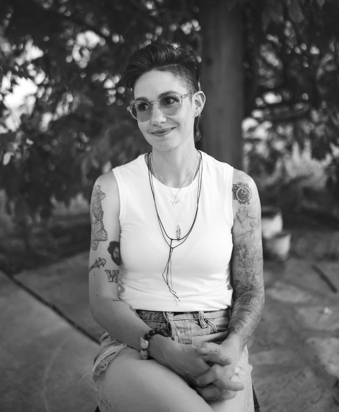 Emily McLoud smiling outdoors under tree in sunglasses, colorful tattoo sleeve