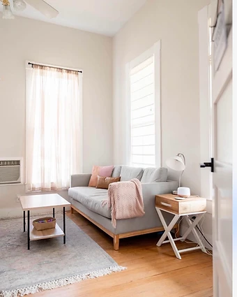 Bright New Braunfels therapy office with a gray sofa, blush accent pillows and a wooden coffee table beside large windows.