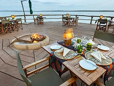 Outdoor wooden deck dining area at Ruzizi Tented Lodge overlooking Lake Ihema, with a set table, fire pit, and canvas-backed chairs