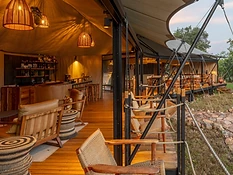 Interior lounge and dining area of Karenge Bush Camp tent, with a bar, woven furniture, and an open deck overlooking the bush at twilight