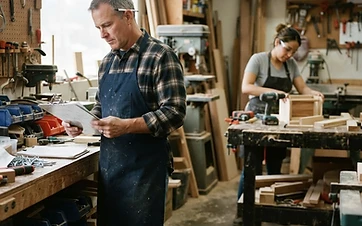 A small-business owner in a workshop reviewing paperwork on a clipboard while an employee works at a bench in the background in natural daylight