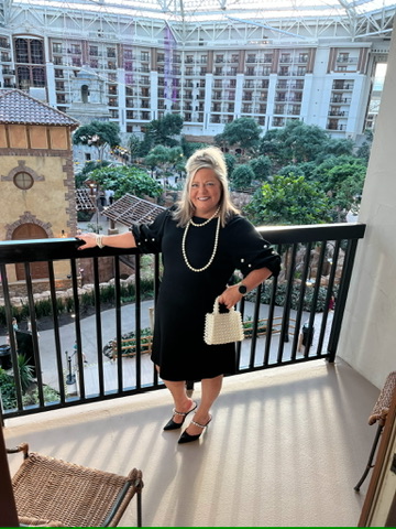 A woman in a black long-sleeved dress with pearl embellishments standing on a resort balcony with a glass atrium and tropical greenery behind her.