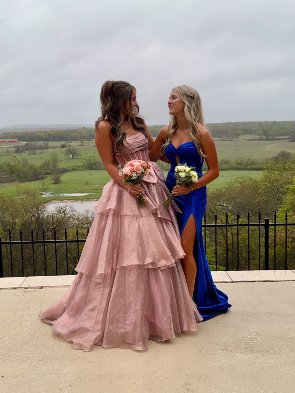 Two young women in formal gowns standing on a terrace overlooking a scenic landscape, one in a dusty-pink tiered dress and the other in a royal blue dress, each holding a floral bouquet.