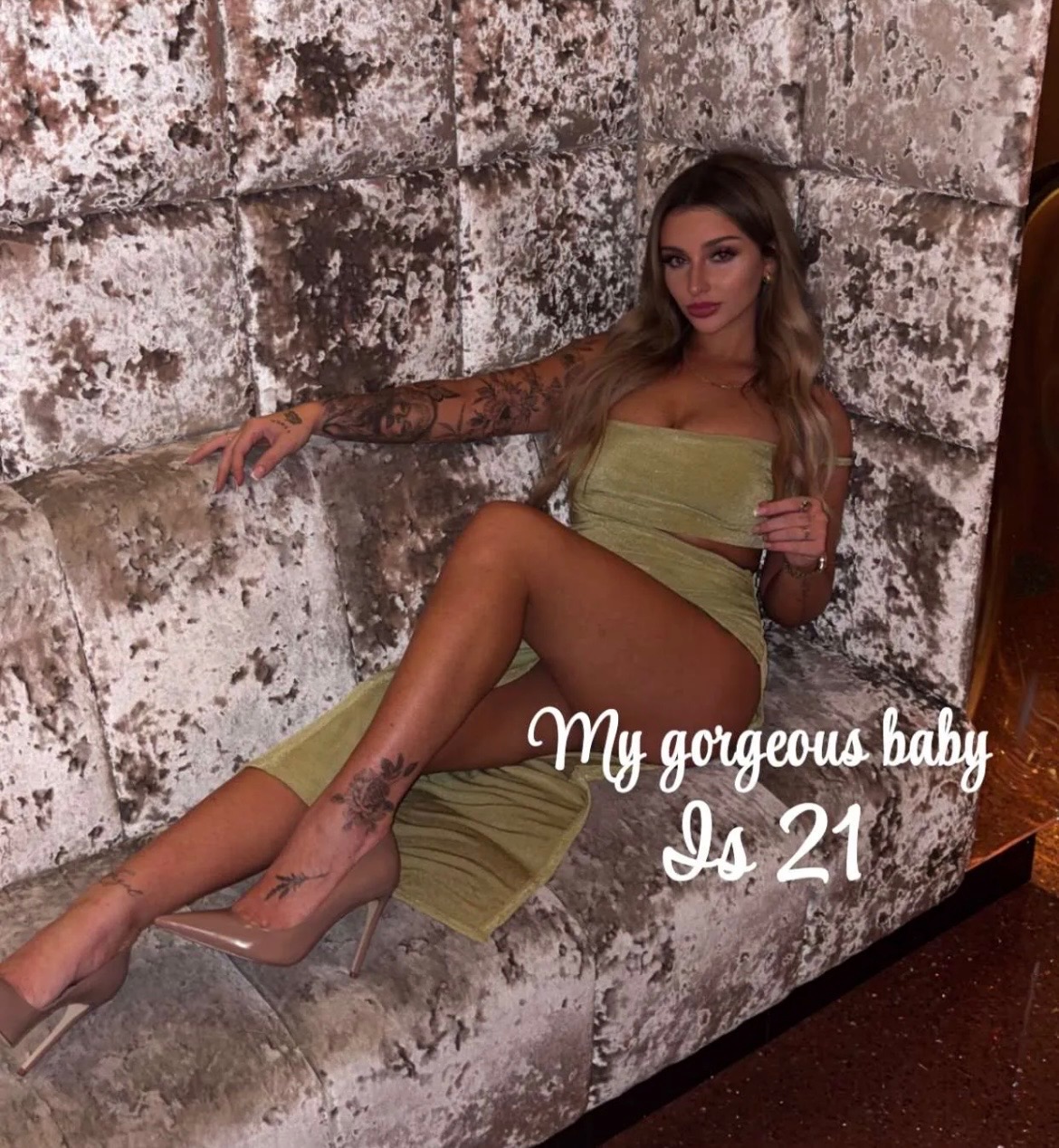 A young woman sitting on a grey velvet couch wearing a metallic olive-green two-piece outfit with tan heels, celebrating a 21st birthday.