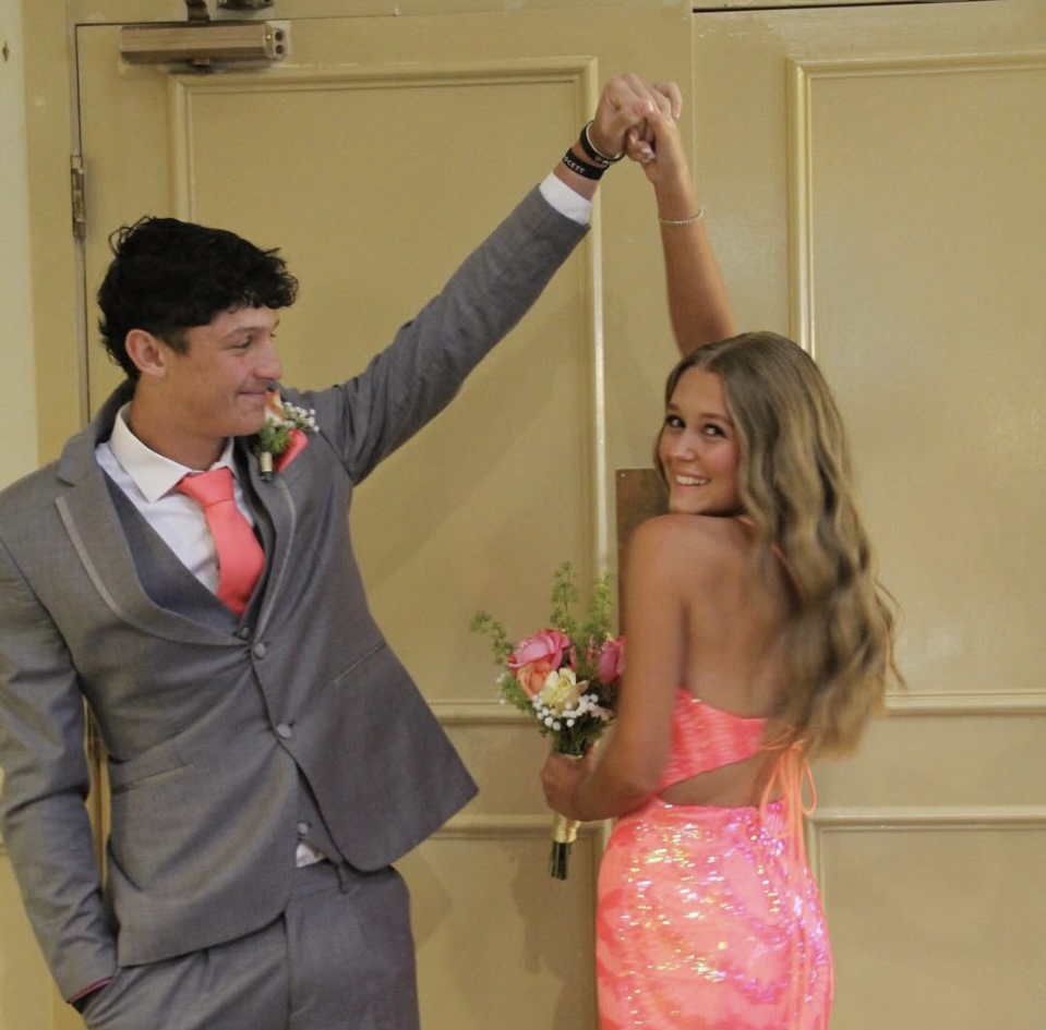 A young couple dancing at an event, the woman in a neon-pink sequined backless dress and the man in a gray suit with a pink tie, holding hands in front of double doors.