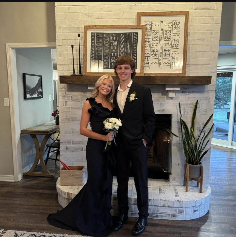 A young couple posing for a formal portrait in front of a white brick fireplace; the woman wears a black one-shoulder evening gown and holds a white rose bouquet, while the man wears a black suit.